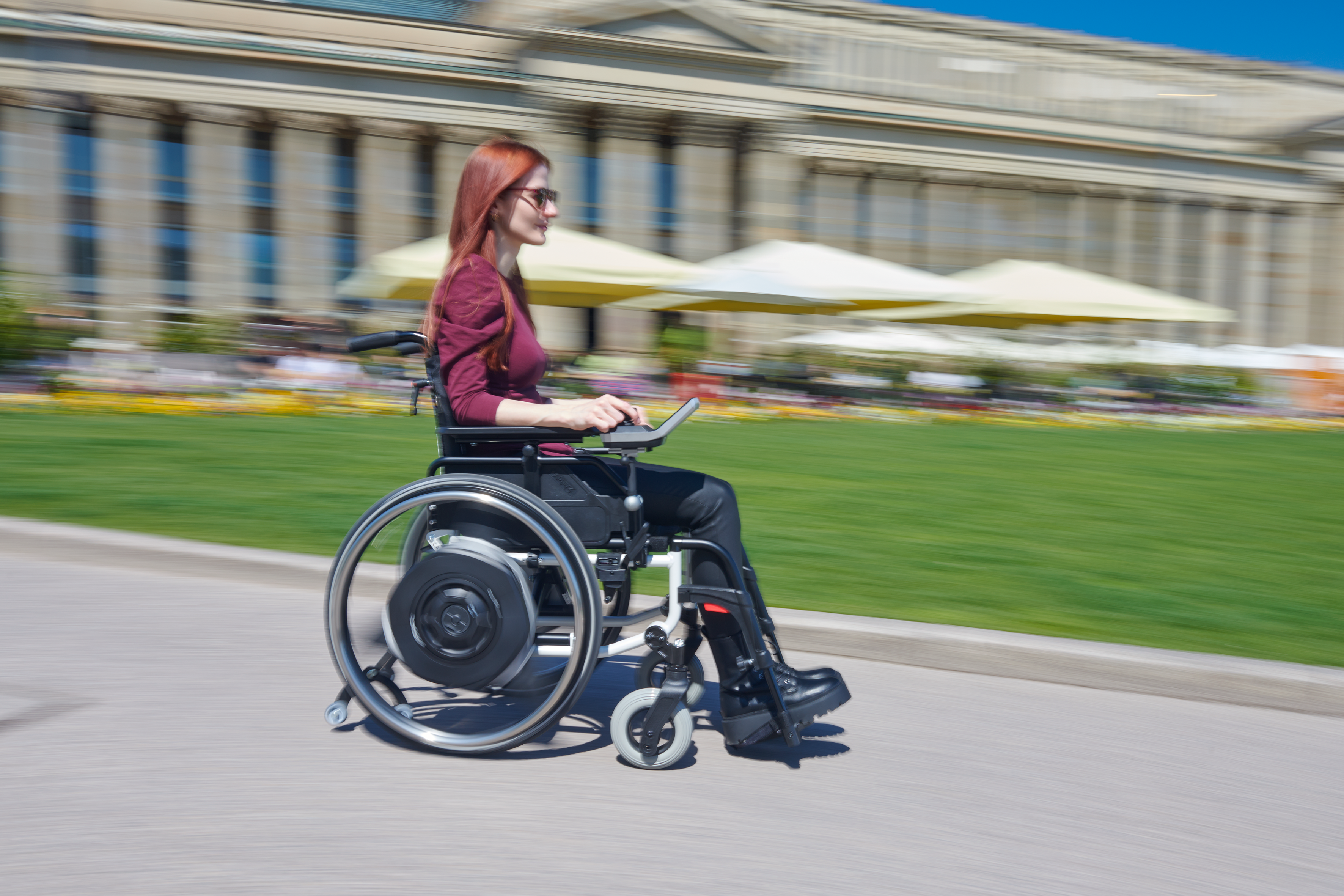 Auf dem Bild ist eine junge Frau in einem manuellen Rollstuhl zu sehen. Angebracht an ihren Rollstuhl ist ein Radnabenantrieb SOLO² der Firma AAT Alber Antriebstechnik GmbH. Das Antriebsrad ersetzt das Standard-Rollstuhlrad und über ein modernes Bediengerät mit Joystick und Farbdisplay wird der Rollstuhl gesteuert. Sie fährt schnell über Pflastersteine in der Innenstadt von Stuttgart. Im Hintergrund ist verschwommen ein Gebäude und die Einkaufsstraße zu sehen. 